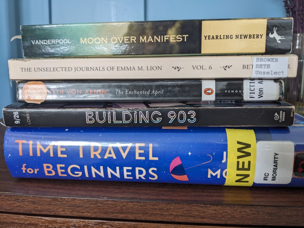 A stack of books, including Moon Over Manifest, The Unselected Journals of Emma M. Lion, The Enchanted April, Building 903, and Time Travel for Beginners