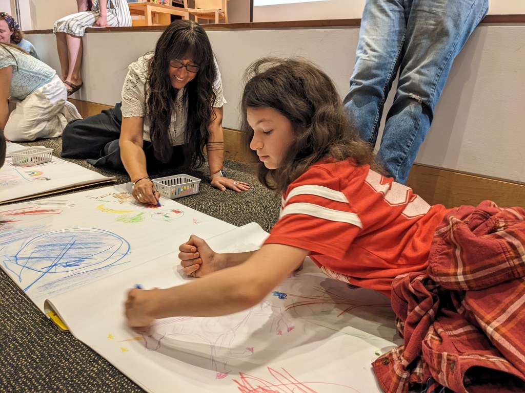 Photo of Juana Martinez-Neal, with long dark hair and glasses, and a light-skinned child in a red t-shirt drawing with beeswax crayons on large paper on the floor