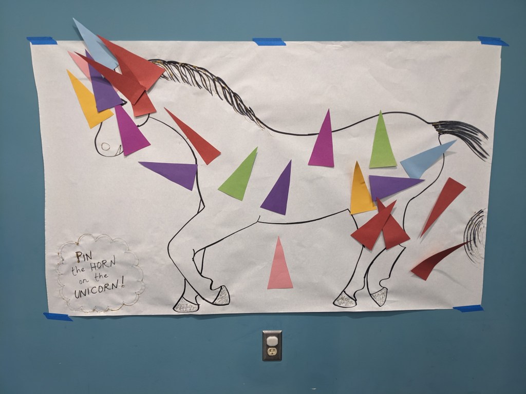 A large piece of white butcher paper with a horse drawn on in black marker, and colorful construction paper horns taped all over
