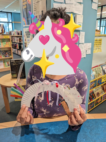 A child in a purple unicorn-print dress holds up her paper plate unicorn craft; her face is blocked out by a sparkle unicorn emoji.